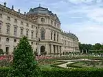 An ornate building on the left side of the picture. In the midground the center of the building projects out, with columns surrounding the main entrance. The right side of the picture is covered in gardens.