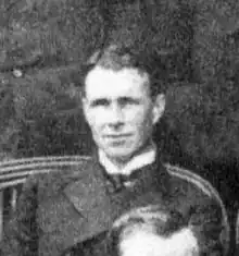  A man, fresh-faced with dark, brushed-back hair, seated among a group. He is wearing a naval officers uniform with a high, stiff collar