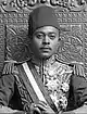 A black-and-white photograph of a man with a dark moustache wearing a turban and a dark jacket and sitting on a throne topped by two metal lions