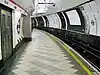 The interior of a building with a rounded roof and walls, a grey door on the left, a railway track on the right, and a man walking in the middle