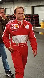 Head, arms and torso of a man in his thirties with his right arm slightly bending. He is wearing a scarlet red racing overalls which displays the Vodaphone, Shell, Bridgestone and Ferrari logos at the front with a white background embroiled on the bottom top.