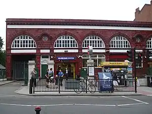 A red-bricked building with eight people standing in front of it and a blue sign reading "BELSIZE PARK STATION" in white letters all under a bright sky