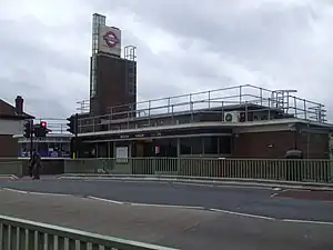 A red-bricked building with a blue sign reading "BOSTON MANOR STATION" and a person in the foreground all under a blue sky with white clouds