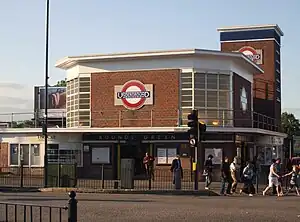 A red-bricked building with a blue sign reading "BOUNDS GREEN" in white letters and several people walking in the foreground all under a blue sky