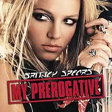 The face of a blonde woman. She is holding a microphone in her hand next to her head, while looking towards the left side of the picture. She is wearing different rings in her fingers. She is wearing a black vest. On the lower part of the image, the words "My Prerogative" are written in red capital letters inside a box of the same color.