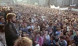 A town square is filled with thousands of people, some holding large banners, looking towards a group of people on a platform in the left foreground. A man with a beard is in the foreground in front of microphones, addressing the crowd.