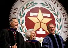 The head and shoulders of three people - an older man, an older woman, and a middle-aged man - wearing formal robes are shown in front of a large circular seal.  On the outer edges of the seal the letters "XAS A...IVERSITY...87..." are visible; an inner band of leaves separates the letters from a block T superimposed with a star.