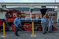 A red steam locomotive with a 4-4-0 wheel arrangement (four leading wheels, four driving wheels, and no trailing wheels) and its tender