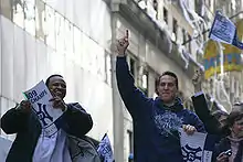 A black man on the left wearing a white T-shirt and dark jacket smiles holding a sign, while a white man on the right wearing a blue sweatshirt which reads "Champions" points upwards. White streamers hang from a building in the background.