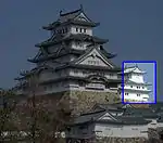 A small three-storied castle tower next to a large five-storied tower. Both have white walls, dark roofs and are built on a platform of unhewn stones.