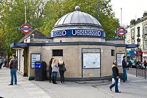 A pink-tiled building topped by a silver dome culminating in a silver ball all surrounded by people under a white sky with green trees in the background