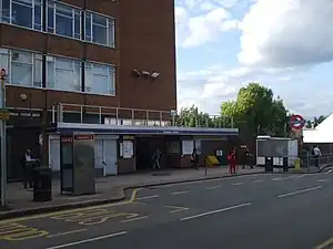 A red-bricked building with a blue sign reading "COLINDALE STATION" in white letters and several people in front all under a blue sky with white clouds