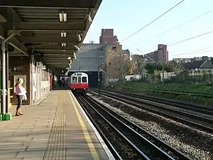 A girl with a pink shirt and blue jeans on a railway platform with a railway track on the right and a white train on it with a red front