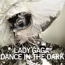 Face of a blond woman, whose platinum blond hair is shaped in a bob cut. She wears a shiny dark colored dress and has a black mask with strings around it on her eyes. The woman tilts her head to the right. Beneath her chin, the words "Lady Gaga" and "Dance in the Dark" are written in white bold font.