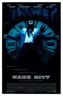 A black poster. Above reads the lines: "Rufus Sewell", "Kiefer Sutherland", "Jennifer Connelly", "and William Hurt". In the center, against a black background, a man wearing a blue jacket is rested against an upright clock with roman numerals as big as him; the setup cast in a blue tint. His arms are outspread, and his head is tilted back with his mouth agape. Behind the man and the clock is a dark city skyline. Below them is the tagline, "They built the city to see what makes us tick. Last night one of us went off." Below the tagline is the film title, "Dark City".