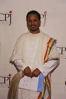 一位擁有棕色皮膚、黑色短髮的中年男性身穿白色長袍,而白袍右邊邊緣是一條由三種顏色組成的線。那些線由右至左分別是綠色、黃色和紅色。該男性手持一束淺黃色毛髮,以及一張證書。該男性正面露笑容。