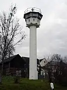 A tall white tower standing near houses and a barn; its base is formed by a slim round column, at the top of which is an octagonal cabin with top-hung windows facing into all directions; there is a searchlight on top of the tower's flat roof, and a railing around the roof's outer edge.