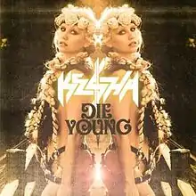 A Caucasian female with blond braids and a floral diadem. She is sparsely clothed in a suit of leather strips with much skin expose. Back facing a mirror, her reflection is symmetrically depicted with the words "Kesha" and "Die Young" appearing in the center.