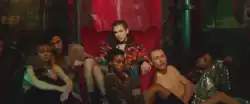 Dua Lipa sitting on a red chair in a warhouse, surrounded by models sitting on the floor.