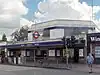 A grey building with a blue sign reading "EALING COMMON STATION" in white letters and two green trees in the background all under a blue sky