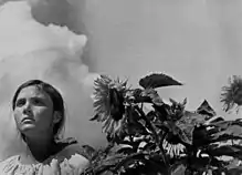 A young woman stands next to a sunflower, against a calm sky