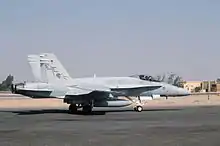 Photo of a modern fighter aircraft moving along tarmac. Sand and buildings are visible in the background.