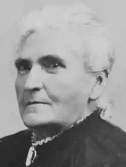 woman with white hair pulled back in black dress with white ruffles showing at collar stares sternly at camera