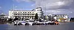 White building in modernist style in the back, waterfront with sailing boats in front