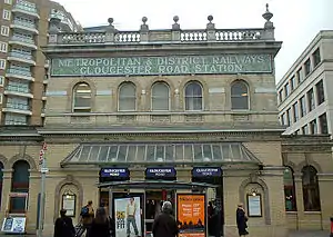 A beige-bricked building with a green sign reading "METROPOLITAN & DISTRICT RAILWAYS GLOUCESTER ROAD STATION" in white letters