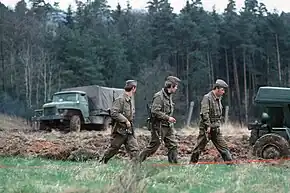 Three uniformed men, armed with assault rifles, walking in a column through a grassy landscape with dense trees in the background. A canvas-sided truck is visible in the left background and part of another vehicle is seen on the right in a stretch of ploughed-up ground.