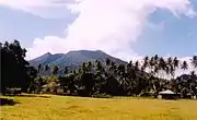 A photograph depicting a blue sky at the top, a grey mountain range in the middle, white clouds in front of the mountain range, and a rocky terrain at the bottom.