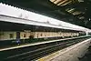 A railway platform with a railway track running from the bottom left corner to the middle right and the legs of several sitting people visible in the bottom right