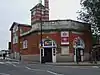 A red-bricked building with a rectangular, blue sign reading "HARROW & WEALDSTONE" in white letters all under a blue sky with white clouds