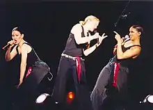 Picture of three woman. The one in the middle has her blonde hair tied in a ponytail. She holds a microphone on her left arm and is leaning towards her right and pointing towards an African American female who's pointing back at her. On the left there's a caucasian female holding a microphone with her left hand.