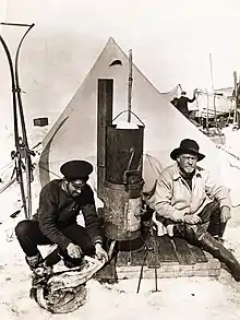  Two men sitting in front of a pointed tent; left-hand man in flat naval cap is skinning a penguin, right-hand man wears a wide-brimmed hat. Between them is a tall stove. Other equipment is visible in the background