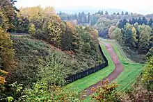 At the base of a wooded slope to the left, there is a black fence running into the distance, flanked on the right by a strip of closely cropped grass, then a strip of bare soil, then another strip of grass, and then a patrol road. Halfway up the wooded slope, a white cross is visible.