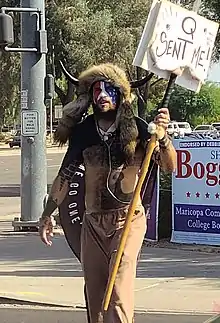 Angeli in his shaman dress on a sidewalk, holding a "Q sent me" sign