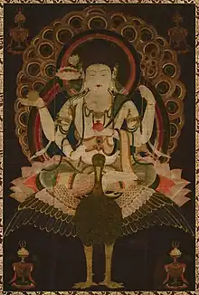 Frontal view of a deity with four arms seated on a bird in lotus position embellished with ornaments.