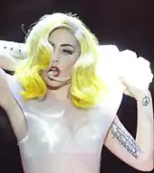 Gaga performing with her hands raised behind her neck. On the left biceps, a line of tattoos is visible.