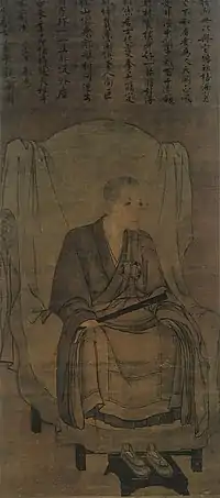 Monk seated on a chair holding a stick-like object in his right hand in three-quarter view. Above the painting there is Chinese text.