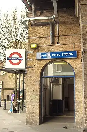 A beige-bricked building with a dark-blue, rectangular sign reading "LATIMER ROAD STATION" in white letters all under a white sky