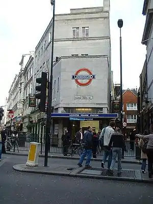 A grey-bricked building with a dark blue, rectangular sign reading "LEICESTER SQUARE STATION" in white letters all under a bright sky