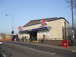 A white-bricked building with a dark blue, rectangular sign reading "LEYTON STATION" in white letters all under a blue sky fading to purple on the horizon