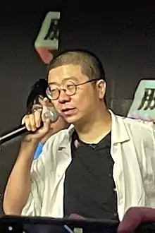 一名男子半身照片，男子穿白色衬衫黑色T恤，手持话筒站在舞台上