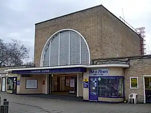 A beige-bricked building with a dark blue, rectangular sign reading "LOUGHTON STATION" in white letters all under a blue sky with white clouds