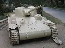 A beige-coloured Locust tank faces the camera, with its gun barrel pointing upwards slightly. Its right-hand track has been damaged and is falling off. The front section of the hull is missing two large loop-shaped handles that other examples possess. It is situated on grassland, with a road passing next to it.