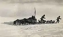  A loaded sledge being pulled across an icy surface by two figures and a team of dogs