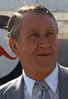 Colour photograph of a Malcolm Fraser aged about fifty, he has a weathered face and greying hair parted on the right. He wears a suit and tie; behind him can be seen part of a large aircraft with a kangaroo logo.