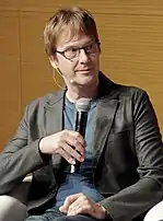 A middle-aged American man holding a microphone wearing glasses, a blue t-shirt, and a black coat