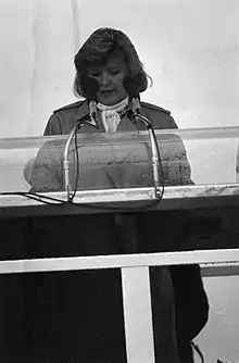 A black-and-white photo of a woman in her fifties making a speech behind a podium and microphone